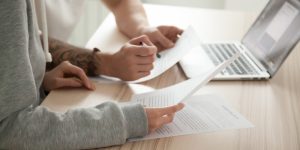 Couple reading legal documents at home with laptop, family considering mortgage loan or insurance, studying contract details, discussing terms and conditions, close up view of hands holding papers