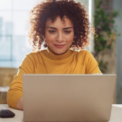 Beautiful Middle Eastern Manager Sitting at a Desk in Creative Office. Young Stylish Female with Curly Hair Using Laptop Computer in Marketing Agency. Colleagues Working in the Background