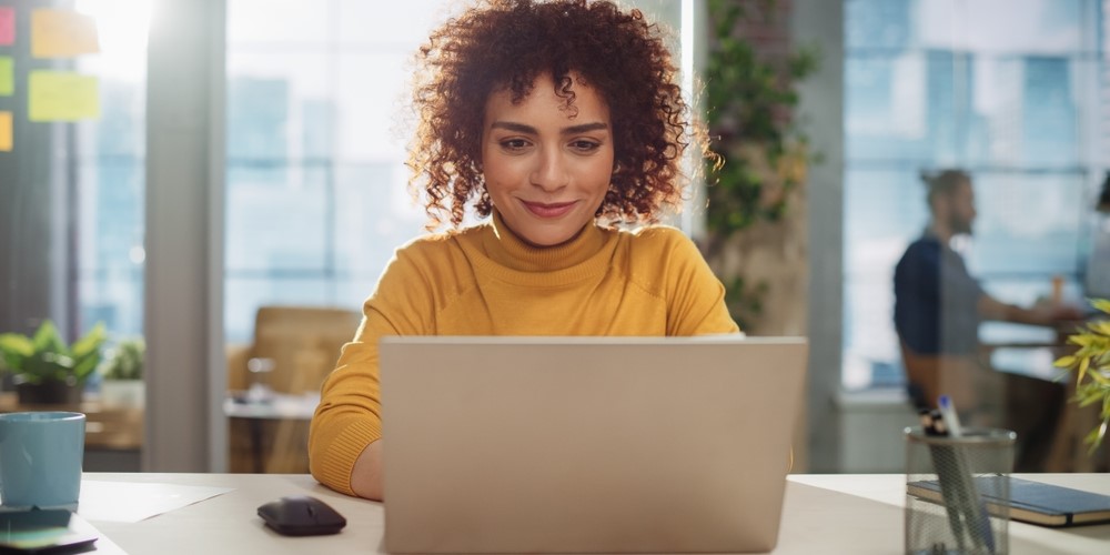 Beautiful Middle Eastern Manager Sitting at a Desk in Creative Office. Young Stylish Female with Curly Hair Using Laptop Computer in Marketing Agency. Colleagues Working in the Background