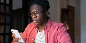 Worried frustrated young student guy sitting at table holding mobile phone, getting bad news via e-mail