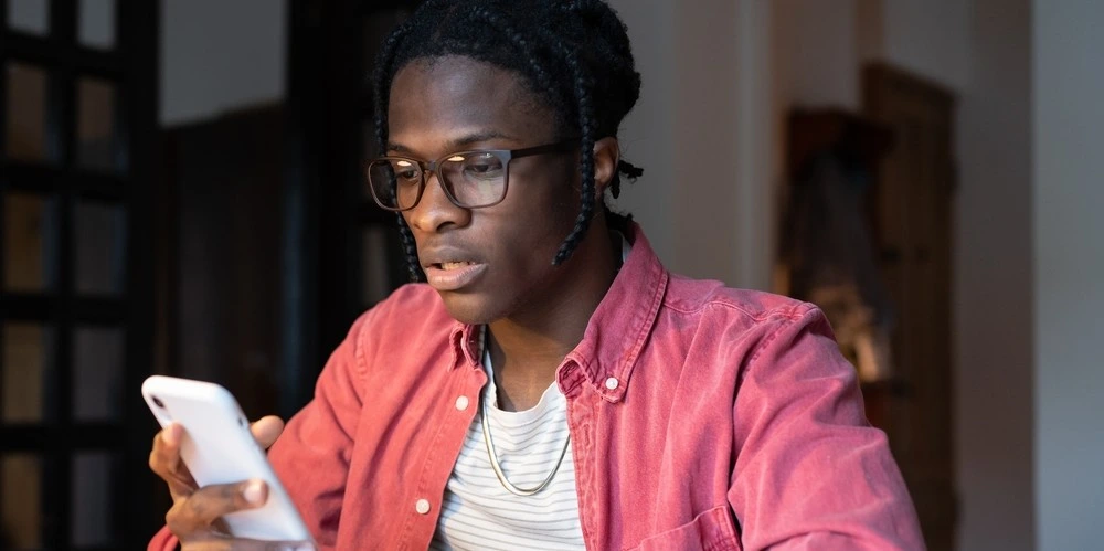 Worried frustrated young student guy sitting at table holding mobile phone, getting bad news via e-mail
