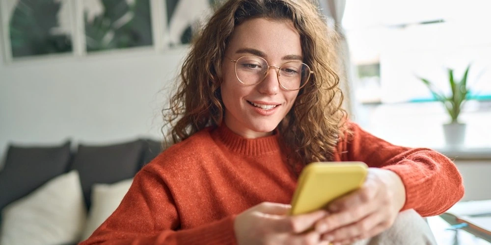 Young smiling woman wearing glasses holding smartphone using cellphone to find out how to claim compensation for a data breach