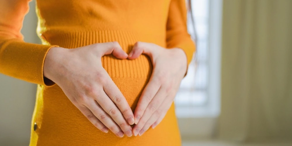 Pregnant woman wearing orange wool dress with hands touching belly in heart shape.